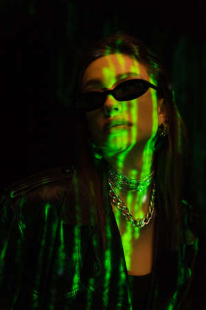 portrait of trendy woman in dark sunglasses and silver necklaces in green neon light isolated on...