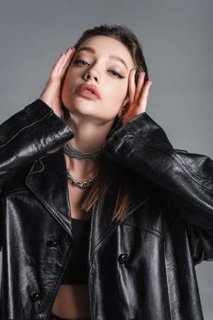 brunette woman in black leather coat and makeup touching face isolated on greyの写真素材