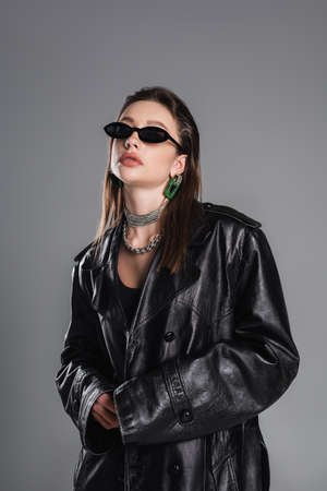 brunette woman in trendy sunglasses and black leather coat posing isolated on greyの写真素材