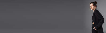 young brunette woman posing with hand in pocket of black trousers isolated on grey, bannerの写真素材