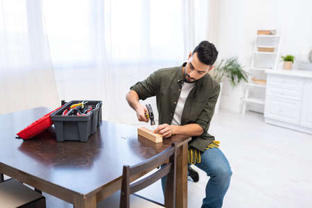 Arabian carpenter holding hammer near nail on wooden board and toolbox at home