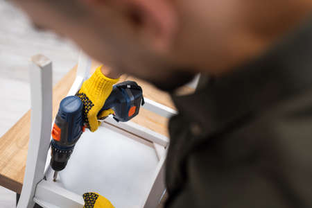 Blurred man in protective gloves fixing chair with electric drill at home