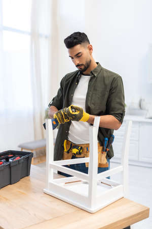 Arabian man in gloves holding electric drill near chair on table at home