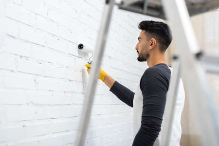 Side view of arabian man coloring brick wall near blurred ladder at home