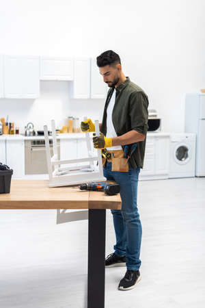 Young muslim craftsman in gloves holding chair near tools at homeの写真素材