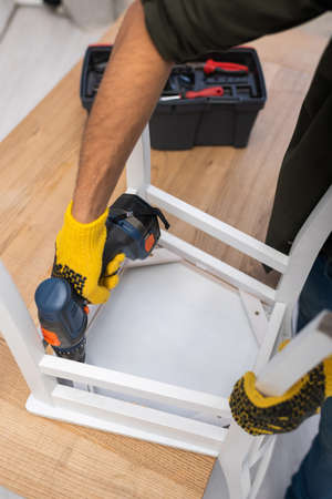 Cropped view of man in gloves repairing chair with electric screwdriver near blurred toolbox at home