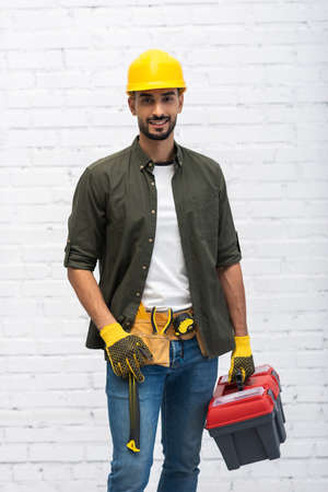 Positive muslim man in hardhat holding toolbox at home