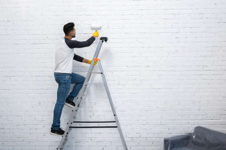 Arabian man in gloves coloring wall while standing on ladder at homeの写真素材
