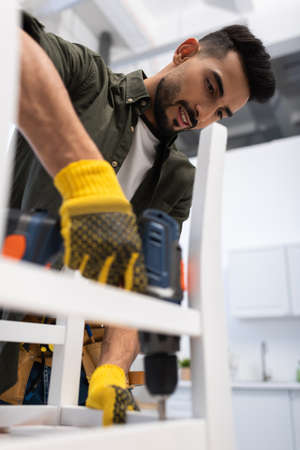 Smiling muslim craftsman fixing chair with blurred electric screwdriver at home