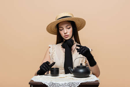 Stylish woman in straw hat and gloves sitting near teapot and cup isolated on beigeの写真素材