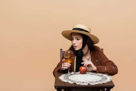 Young woman in sun hat touching wine glass and apple on table isolated on beigeの写真素材