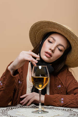 Portrait of woman in sun hat touching glass of wine on table isolated on beigeの写真素材