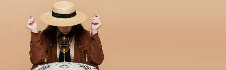 Trendy young woman in sun hat sitting near wine glass of table isolated on beige, bannerの写真素材