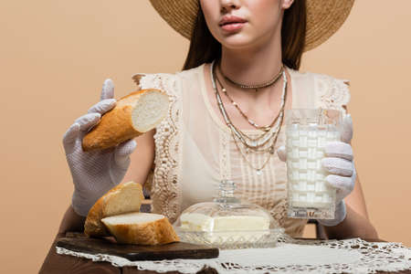 Cropped view of young woman in gloves holding bread and milk near butter isolated on beigeの写真素材