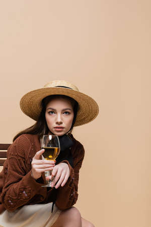 Trendy woman holding glass of wine and looking at camera isolated on beigeの写真素材