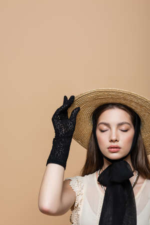 Stylish woman in vintage clothes touching straw hat isolated on beige