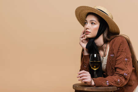 Fashionable model in straw hat looking away near glass of wine on chair isolated on beigeの写真素材