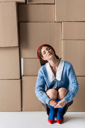 Stylish woman in eyeglasses and beret closing eyes near cardboard boxes on white backgroundの写真素材