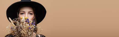 Brunette woman in fedora hat looking at camera near dry plants isolated on beige, bannerの写真素材