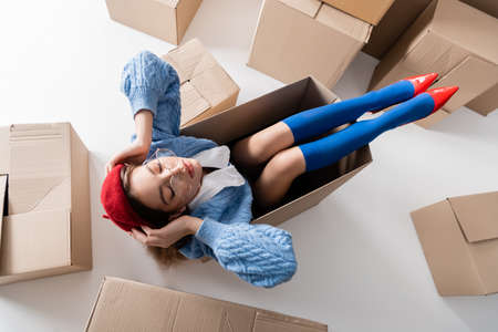 Top view of stylish woman in beret and eyeglasses sitting in package near cardboard boxes on white backgroundの写真素材