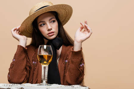 Stylish woman in straw hat looking away near glass of wine on table isolated on beigeの写真素材