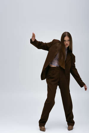 full length of stylish teenage model in brown high-quality suit gesturing on greyの写真素材