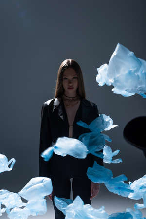 brunette model in black suit posing near flying blue plastic bags on dark grey