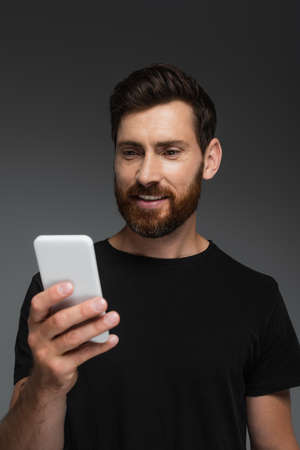 pleased and bearded man in black t-shirt using mobile phone isolated on grey