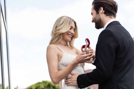 Low angle view of smiling man holding engagement ring near girlfriend in dress on terrace