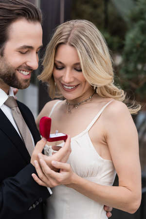 Smiling man in suit holding box with engagement ring near girlfriend outdoors