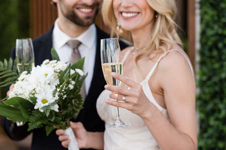 Cropped view of glass of champagne in hand of blurred bride and groom outdoorsの写真素材