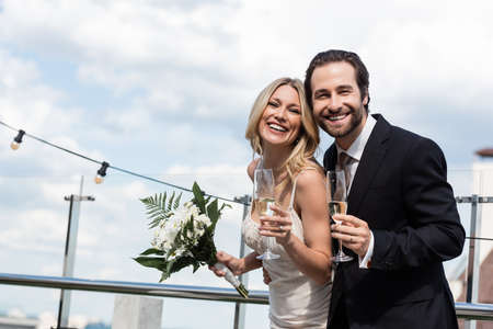 Cheerful newlyweds with bouquet and champagne looking at camera on terraceの写真素材