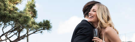 Positive bride with glass of champagne standing near groom outdoors, bannerの写真素材