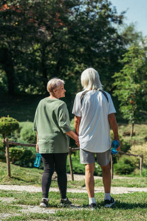 happy retired woman in sportswear looking at husband with sport bottle and holding hands in park