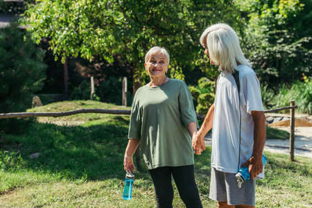 happy retired couple in sportswear with sport bottles holding hands in park