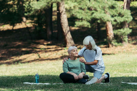 cheerful senior man hugging wife with grey hair sitting on fitness mat in green parkの写真素材
