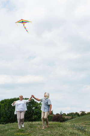 happy senior couple in casual clothes holding hands while playing with kite on green hillの写真素材
