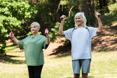 cheerful senior couple in sportswear exercising with dumbbells in green parkの写真素材