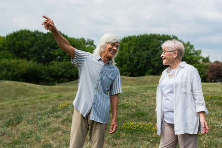 happy senior man in glasses pointing away near wife with grey hair standing on green hillの写真素材
