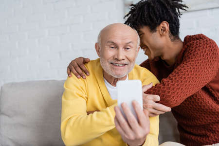 Smiling african american grandson hugging grandparent with smartphone at homeの写真素材
