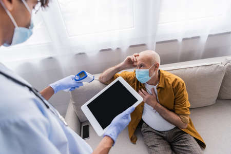 Elderly man in medical mask sitting near blurred african american nurse with pyrometer and digital...