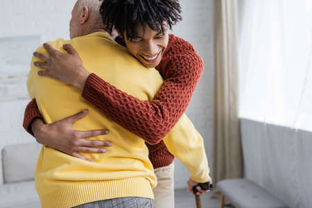 Happy african american man hugging senior grandparent with walking cane at homeの写真素材