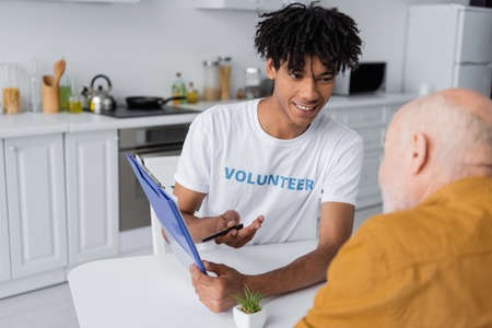 Cheerful african american volunteer pointing at clipboard near blurred senior man in kitchenの写真素材