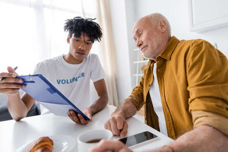 African american volunteer holding clipboard near senior man with cup and smartphone at home