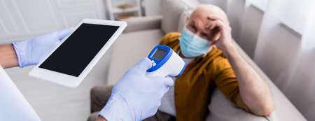 African american nurse in latex gloves holding pyrometer and digital tablet near blurred senior man in medical mask at home, bannerの写真素材
