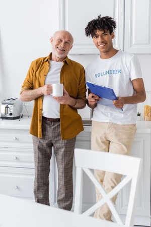 Smiling african american volunteer with clipboard and pensioner with cup looking at camera in kitchenの写真素材