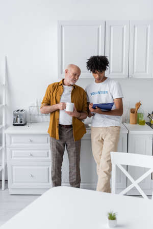 Elderly man holding cup near african american volunteer with clipboard in kitchen