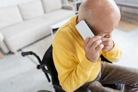 High angle view of displeased pensioner talking on smartphone in wheelchair at home