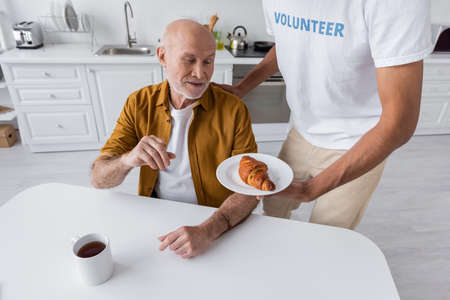 African american volunteer holding croissant near senior man and cup of tea at home