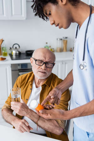 African american nurse pointing at pills near senior patient with glass of water at homeの写真素材